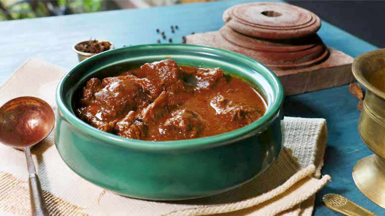 Rampuri Mutton Curry in a emerald green ceramic bowl lying on a cream and gold table mat next to a copper spoon. On a blue table top - Taar Gosht Recipe